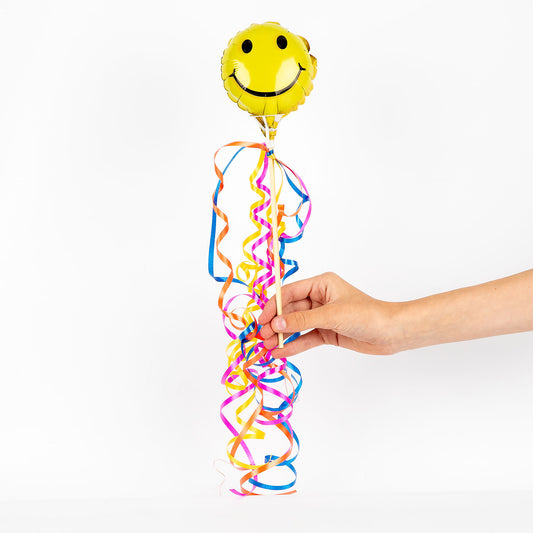 Hand holding a yellow smiley face balloon with colorful ribbons against a white background