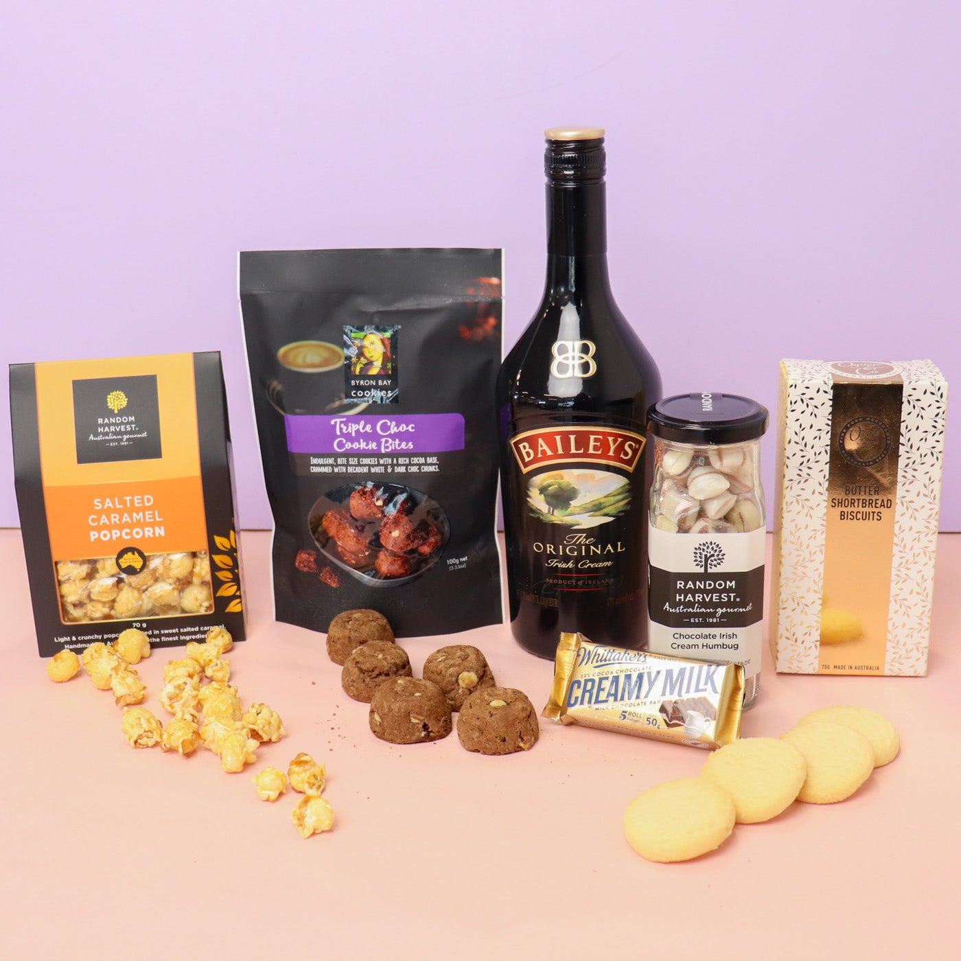 Assorted snacks and a bottle of Baileys on a pink surface with a purple background