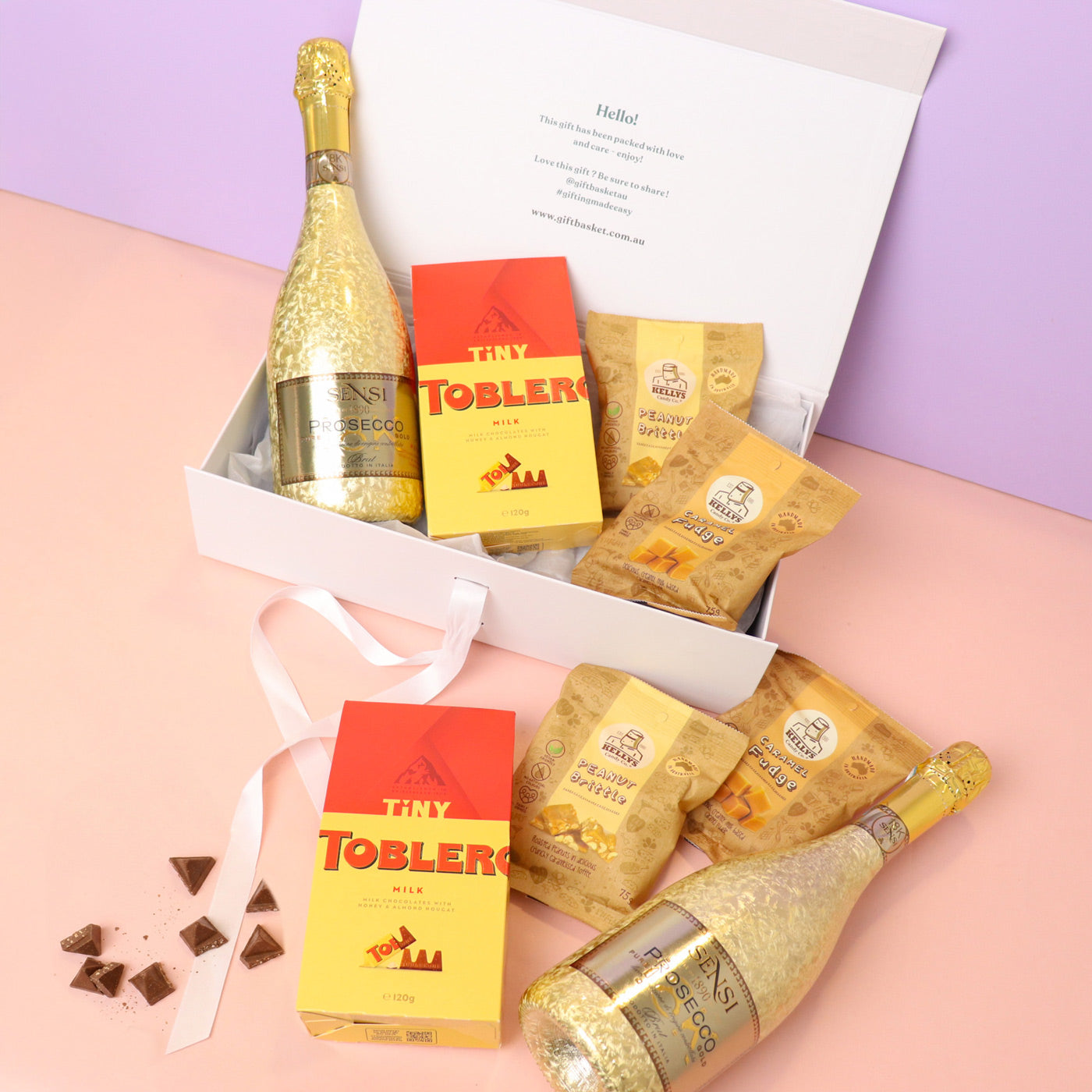 Gift set with gold bottles, Toblerone chocolate, and cookies on a pink and purple background