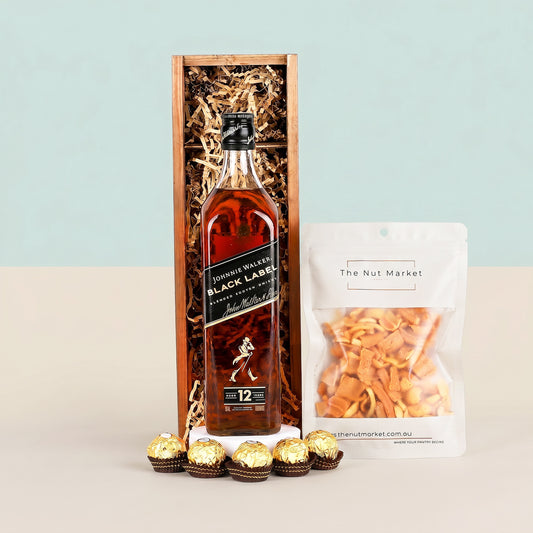 Johnnie Walker Black Label whisky bottle with a pack of Ferrero Rocher chocolates and a bag of The Nut Market snacks on a light blue background.