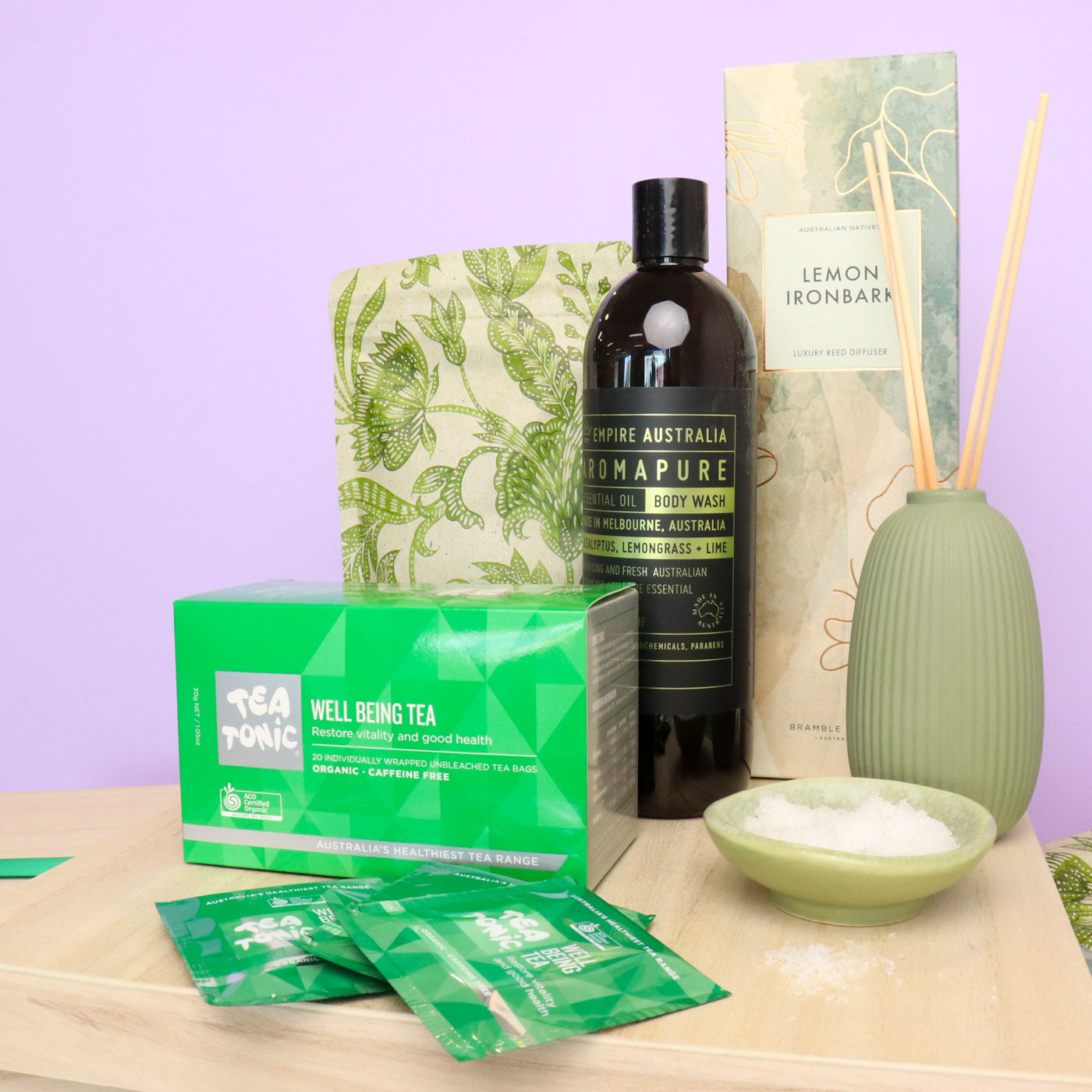 Collection of wellness products including a bottle, tea packages, and a diffuser on a wooden surface with a purple background.