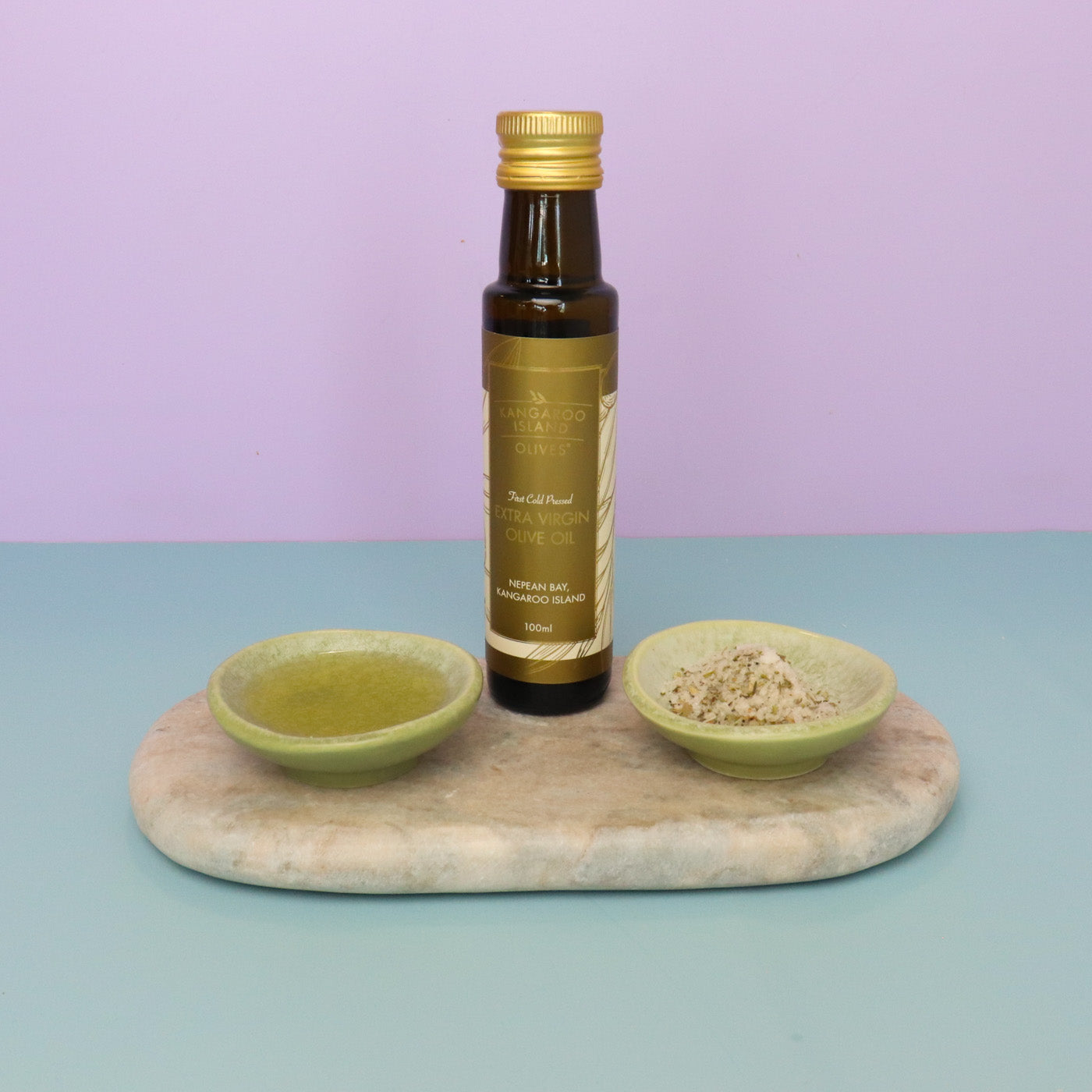 Bottle of olive oil with two small bowls on a marble slab against a pastel purple background