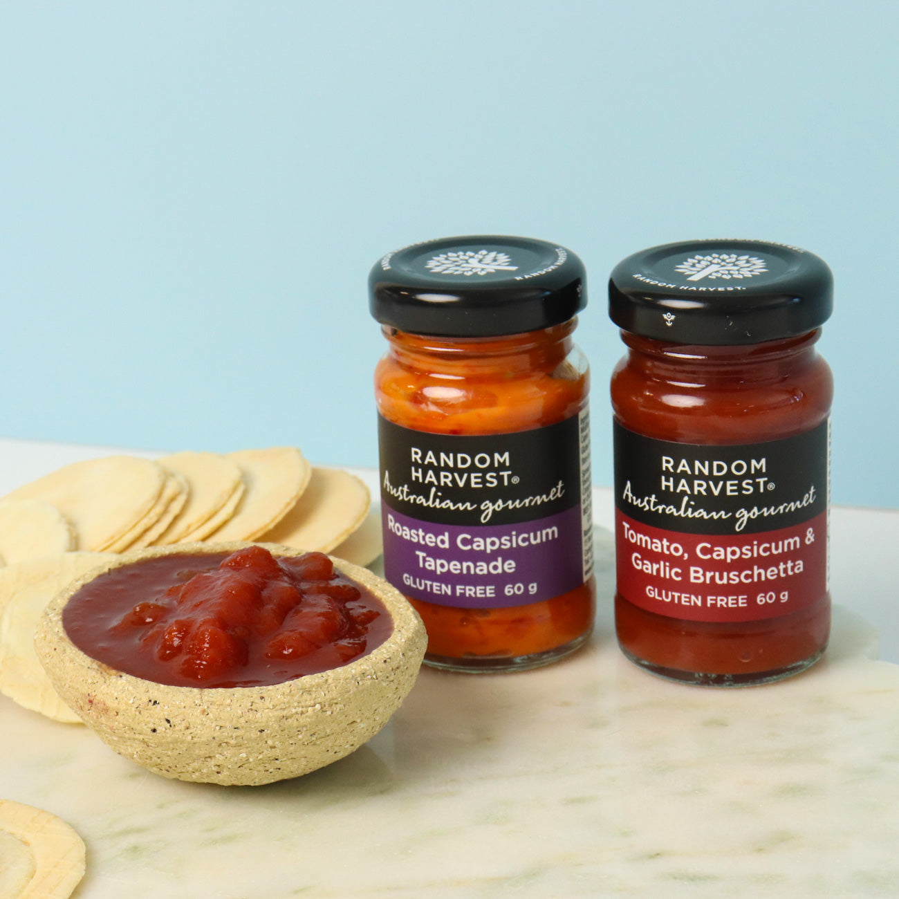 Two jars of Random Harvest tapenade on a marble surface with a light blue background