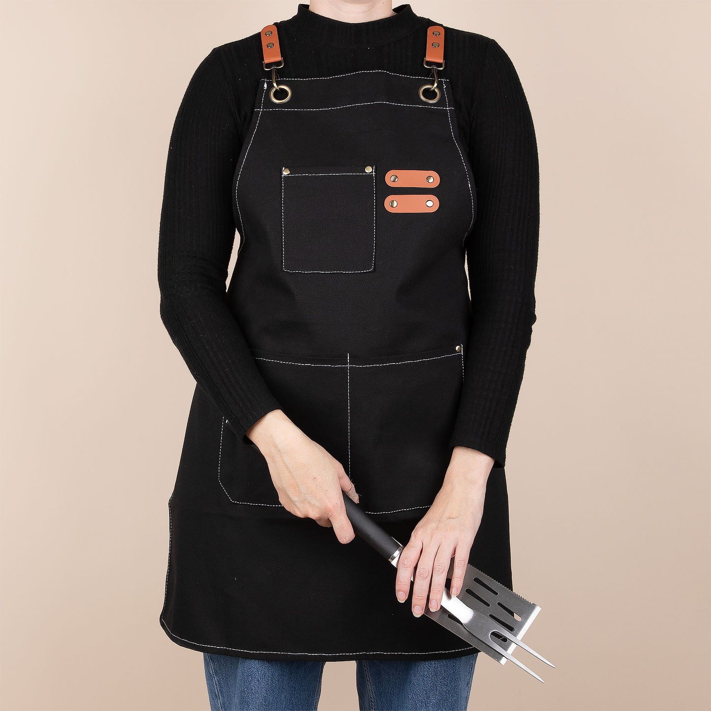 Person wearing a black apron with brown straps holding BBQ utensils on a beige background