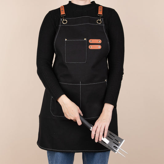 Person wearing a black apron with brown straps holding BBQ utensils on a beige background