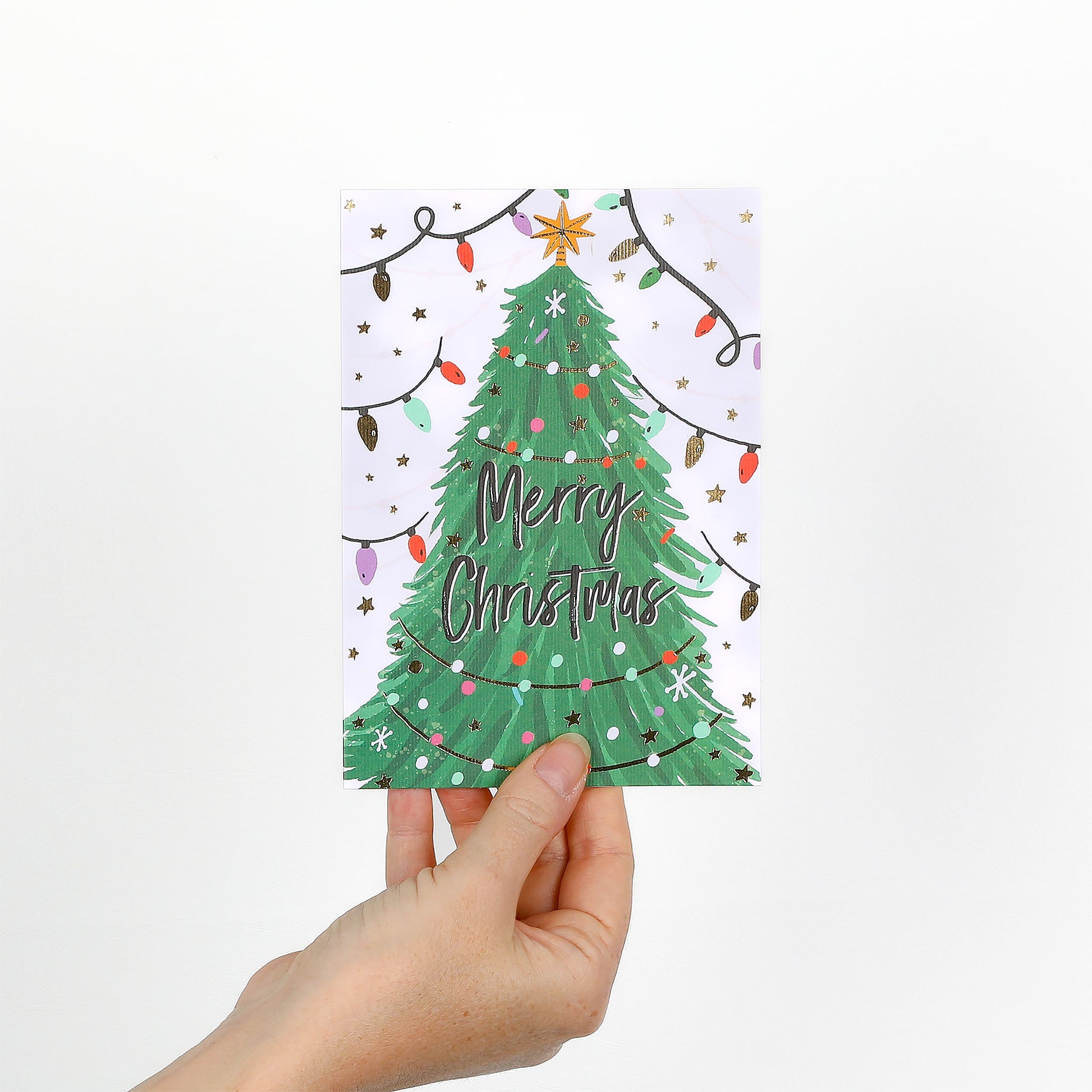 Hand holding a Christmas card with a green tree and 'Merry Christmas' text on a white background