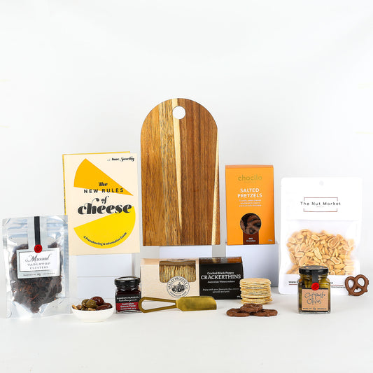 Collection of gourmet food items including a wooden cutting board, cheese book, and various snacks on a white background.
