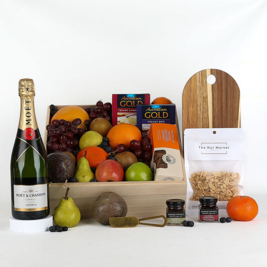 Gift basket with Moët & Chandon champagne, serving board, fruits, and snacks on a white background