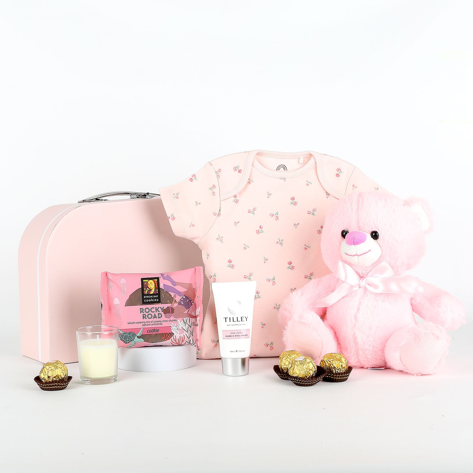 Pink baby outfit with teddy bear, chocolates, and skincare product on a white background