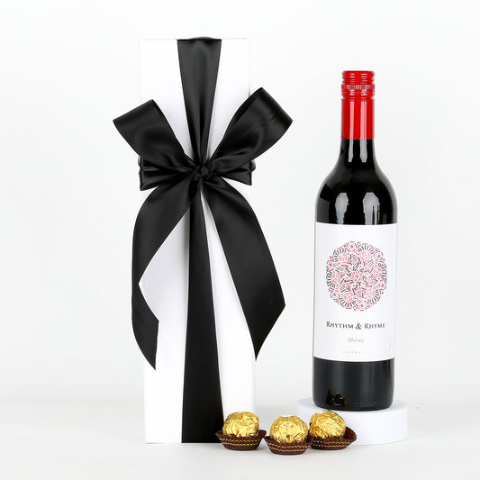 Bottle of red wine with a black ribbon, white box, and gold chocolates on a white background