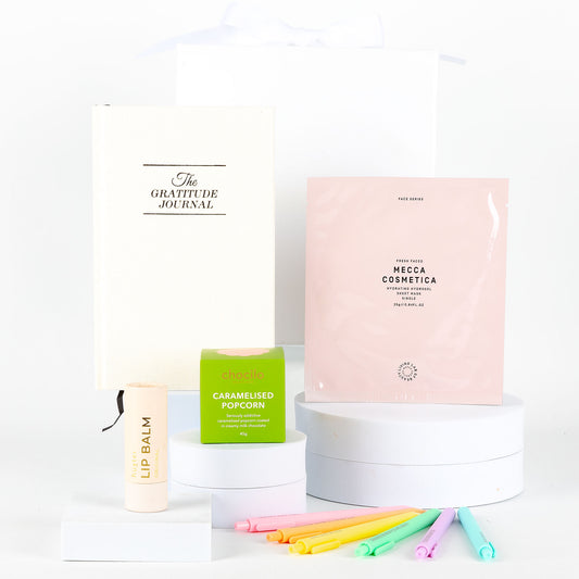 Collection includes a journal, hydrating face mask, lip balm, caramel popcorn, and colourful pens on a white background.